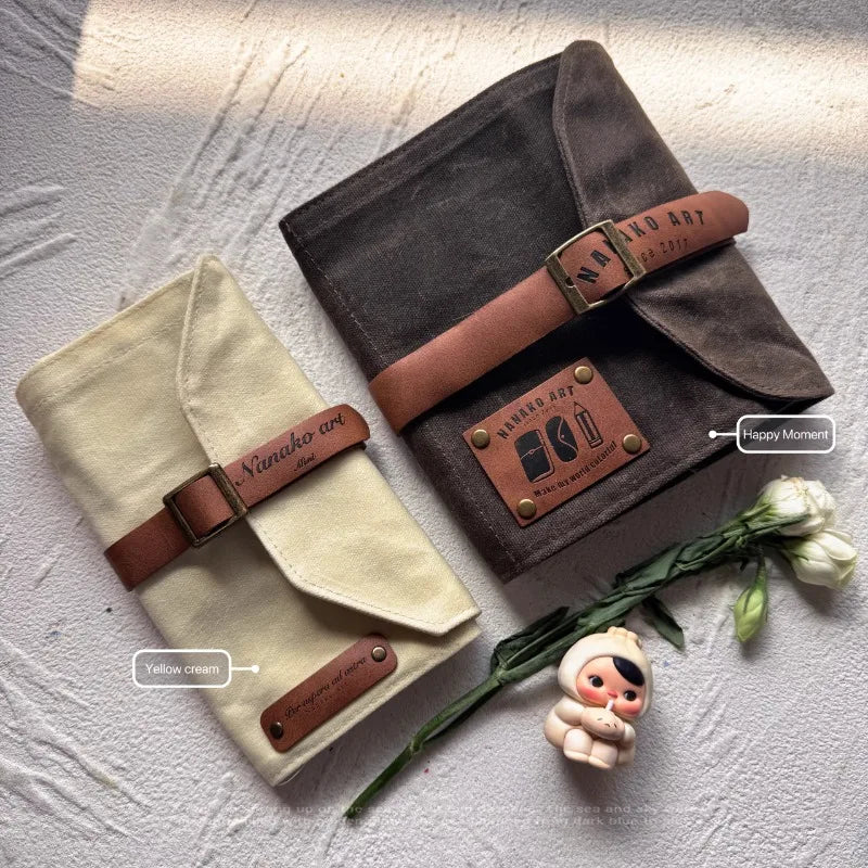 Two fabric pencil cases wraps with brown straps on a textured surface with a small figurine and flowers.